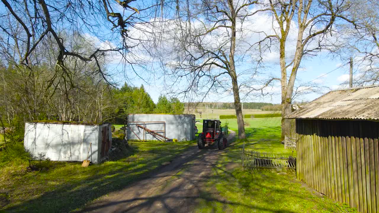 Aerial drone footage flying behind an old retro tractor that is wokring in a sunny countryside rural garden between sheds and on a muddy rural road during a sunny day with blue sky and forest visible.