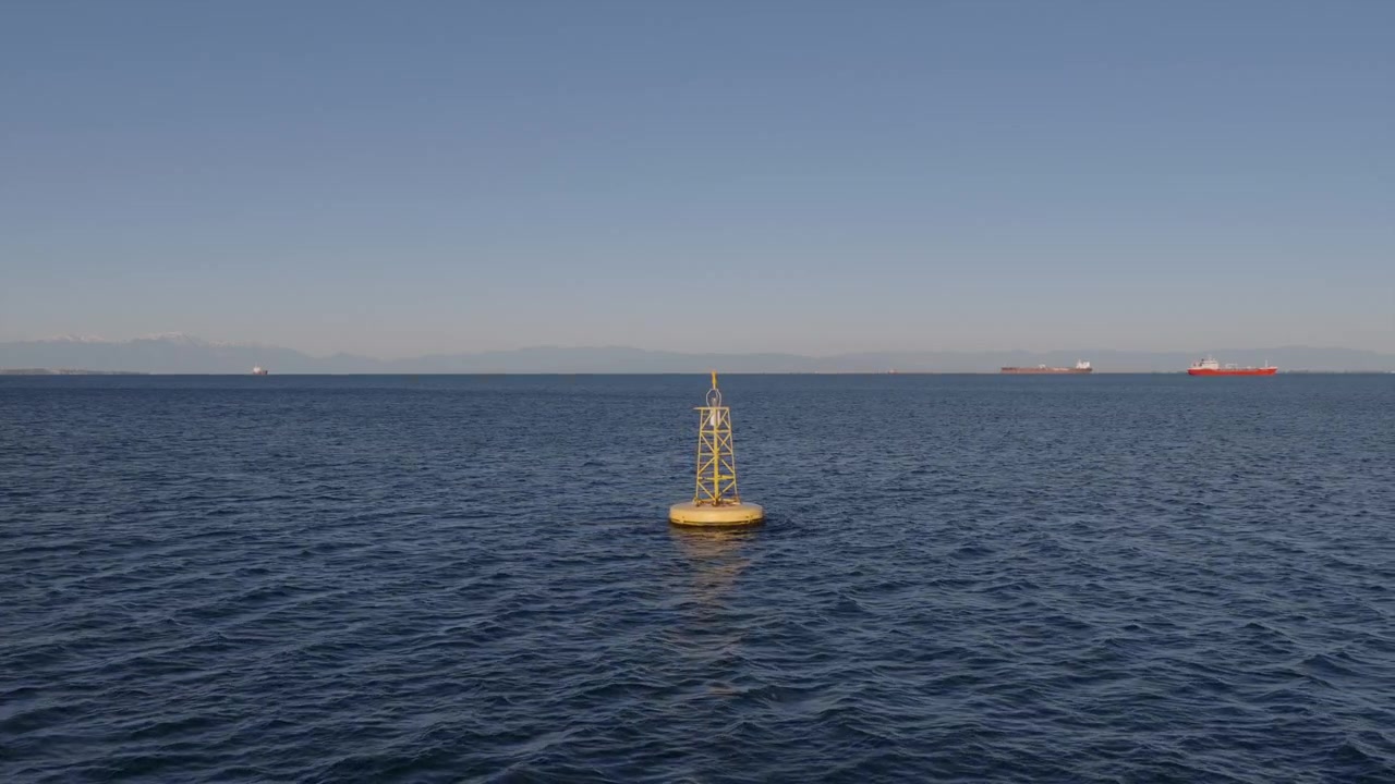 Una boya de navegación amarilla flota en las aguas del golfo cerca de Tesalónica, con cargueros distantes en el horizonte.