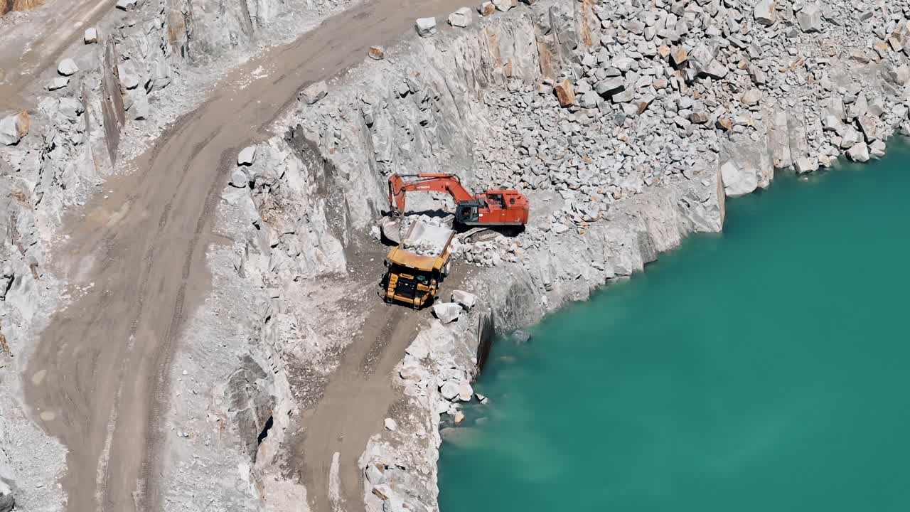 excavadora llenando un camión de basura en una cantera por un lago azul sereno - aéreo