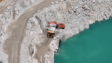 Excavator-filling-a-dump-truck-in-a-quarry-by-a-serene-blue-lake---aerial