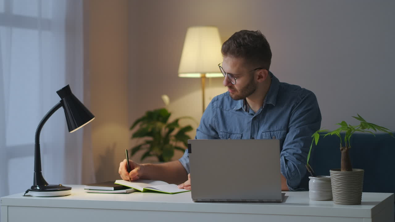 adult man is viewing lecture on display of laptop and making notes in notebook learning online looking for information in internet e-learning and self-education
