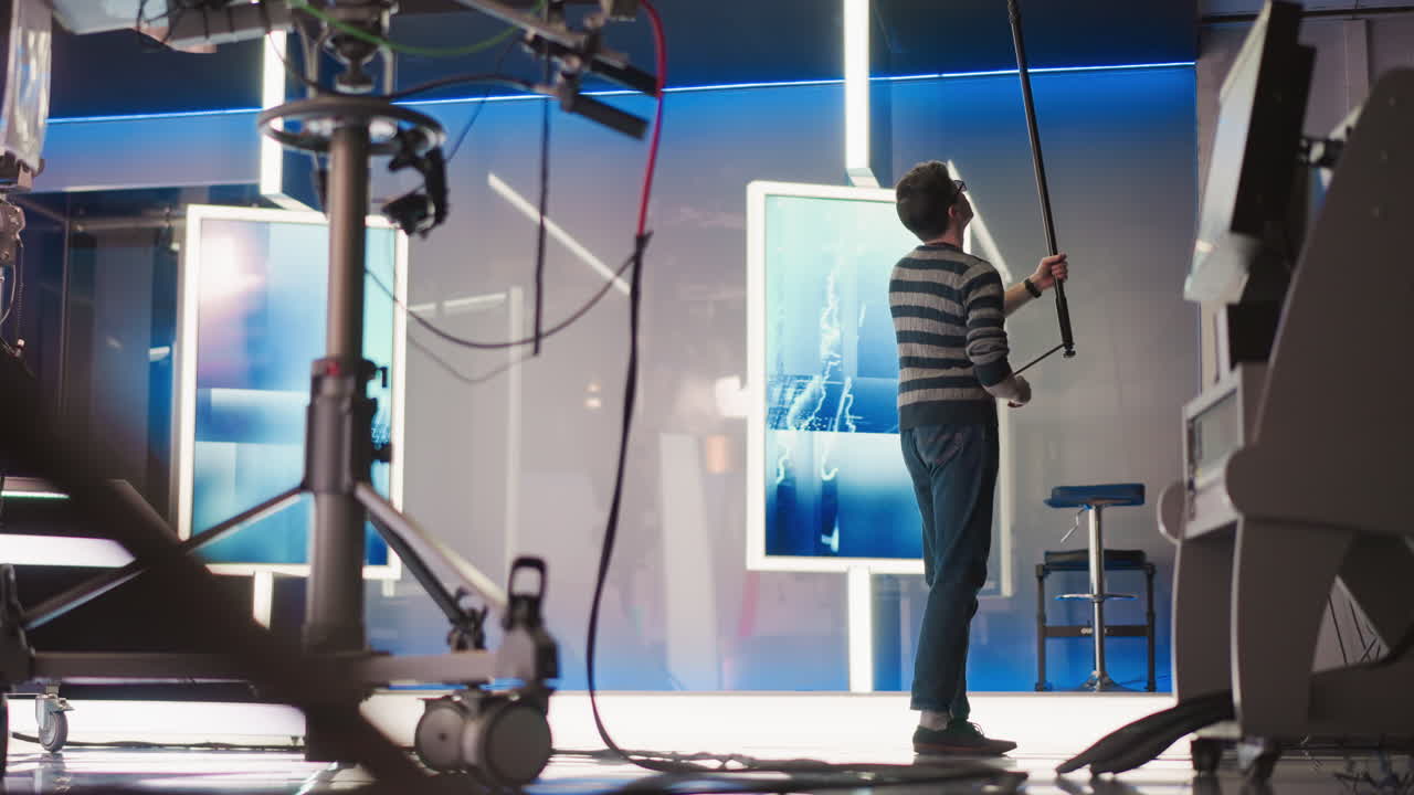 Man adjusting boom microphone in professional video studio, focusing on sound recording while surrounded by high-tech equipment and neon lights, preparing for filming process
