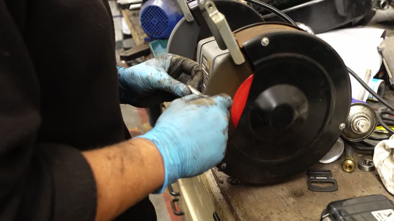 Close-up of a mechanic using a bench grinder to polish a small metal part in a cluttered workshop, wearing protective gloves. Manual metalwork in a professional repair environment