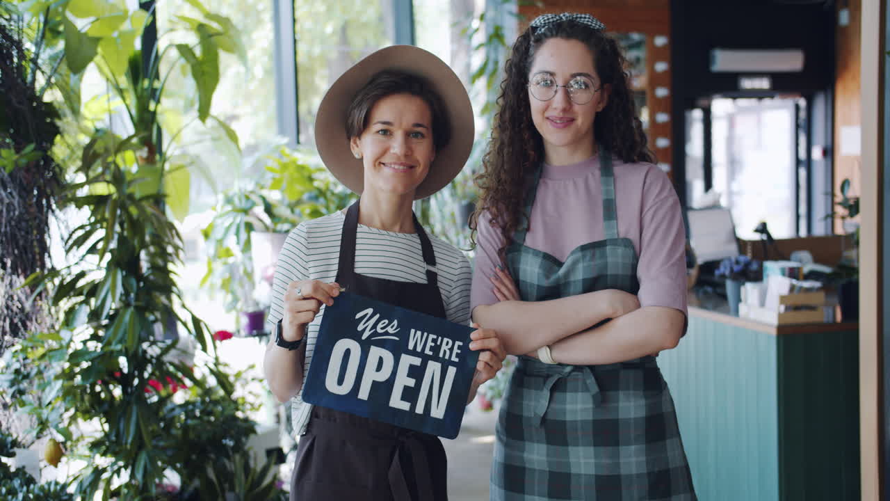 Due fioriste, proprietarie di un negozio, che sorridono e mostrano un cartello "aperto"