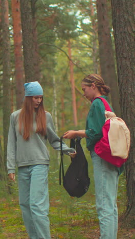 dos mujeres jóvenes en una escena de bosque tranquilo, una vestida con una camisa azul con una bolsa roja ayuda a una amiga con un pañuelo azul a ponerse su bolsa negra, rodeada de árboles altos y vegetación exuberante