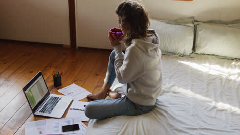 mixed race woman working at home, sitting on bed using laptop and drinking tea in cottage