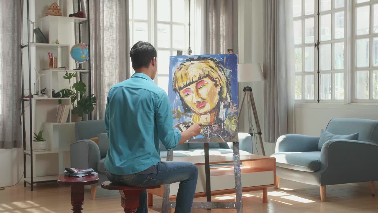 Asian Artist Man Sitting On Round Wooden Chair Without Backrest Holding Paintbrush Mixed Colour And Thinking Before Painting A Girl'S On The Canvas