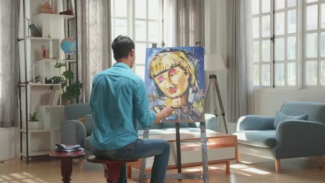 asian artist man sitting on round wooden chair without backrest holding paintbrush mixed colour and thinking before painting a girl's on the canvas