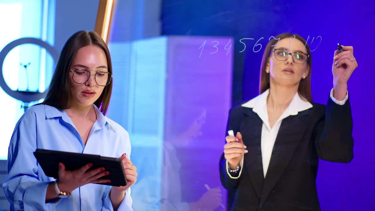 Long-haired brunette in glasses and blue shirt uses a tablet. Lady in black jacket writes on the glass board.