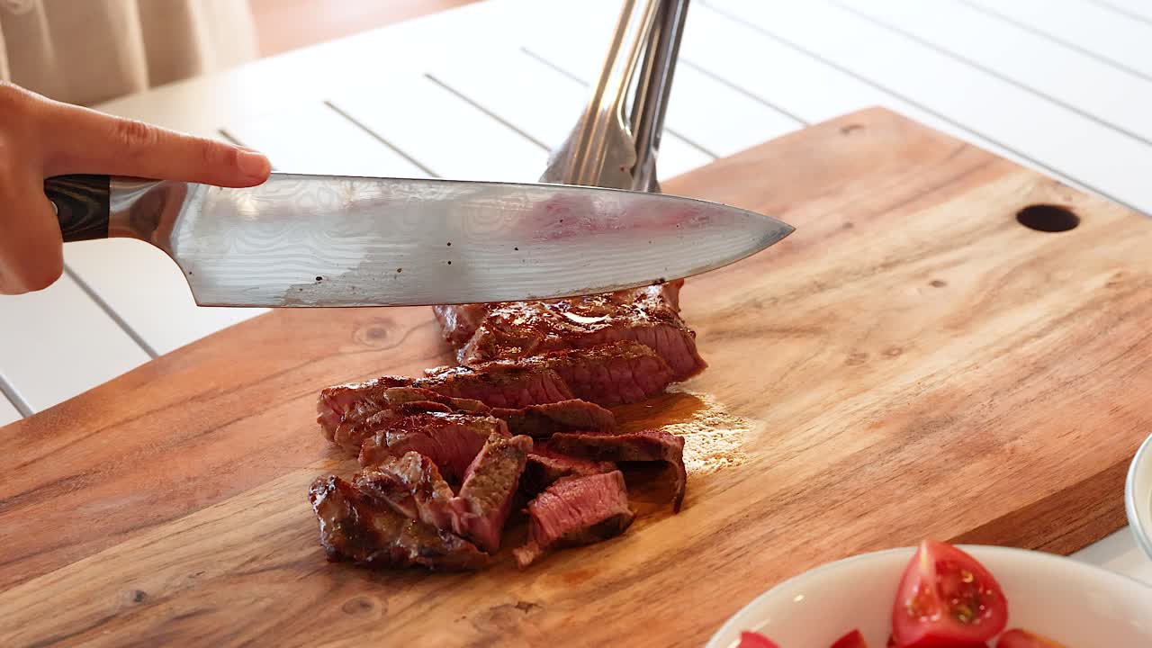 A person slices a cooked beef steak on a wooden board in a well-lit kitchen setting