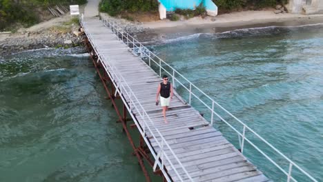 Drohnenansicht-Eines-Jungen-Mannes-Mit-Rucksack-Und-Sonnenbrille,-Der-Auf-Einem-Holzsteg-Am-Robinson-Beach-An-Der-Bulgarischen-Schwarzmeerküste-Läuft