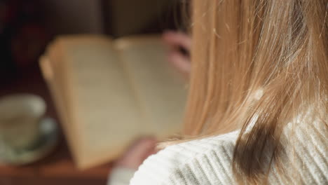 close up rear view of lady with golden hair focused on reading book while flipping pages as sunlight gently highlights her hair and ceramic cup rests on wooden table in cozy indoor space