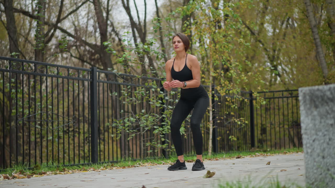 Rotational view of athletic woman squatting down for training outdoors, close to iron fence, dry trees, concrete bench, and well-maintained pathway in the background