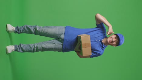 full body of asian male courier in blue uniform showing call me gesture and smiling while delivering a carton on green screen background in the studio