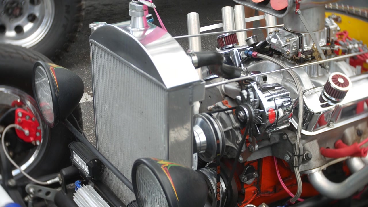 Engine parts exposed on a custom hot rod at a car show. Close-up view shows chrome radiator, belts, headlights, racing tires, and polished mechanical detail