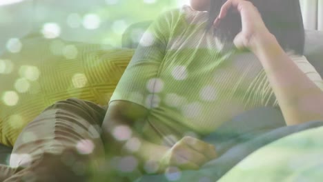 Animation-of-light-spots-over-biracial-woman-sitting-on-sofa