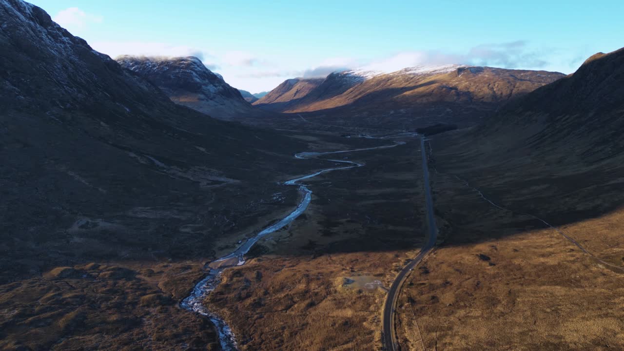 Valle de Glencoe con un río serpenteante y una carretera entre montañas escarpadas, luz del día, vista aérea.