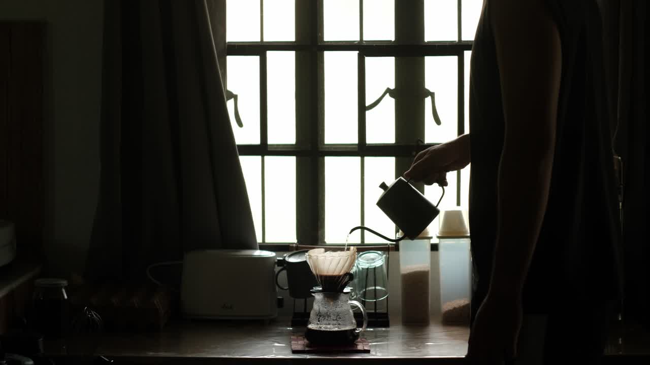Medium static shot of the silhouette of young man pouring hot water through a gooseneck kettle into coffee pour-over setup in apartment kitchen setting