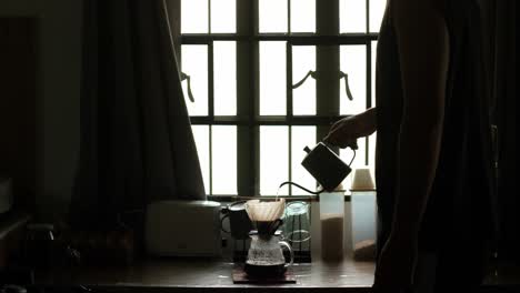 medium static shot of the silhouette of young man pouring hot water through a gooseneck kettle into coffee pour-over setup in apartment kitchen setting