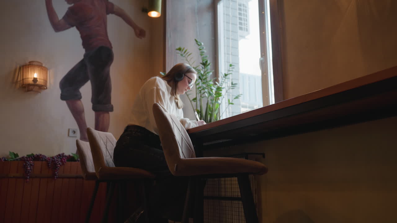 writer wearing headset sits by window jotting notes in notepad with indoor plant and wall mural behind, hand resting on ear as ambient light from outside brightens cozy modern interior space