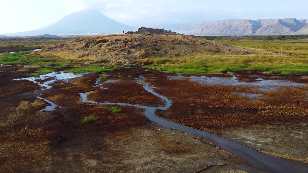 An incredible drone shot of the stunning landscape of Lake Natron with people standing on a hill top with Ol Doinyo Lengai volcano in the background in Tanzania in North Africa