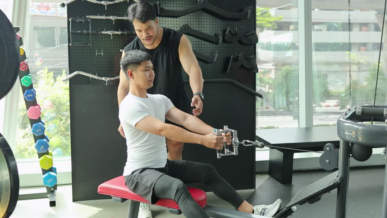 A man exercises on a cable machine with guidance from a trainer in a well-lit gym