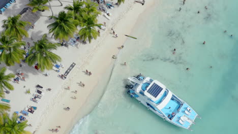 local tourists walk along a white sand beach, a luxury party yacht anchored on the shore, aerial top down