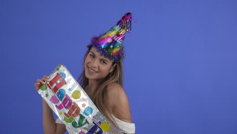 a pretty brunette smiling while holding a happy birthday sign and wearing a party hat, isolated on a blue studio background