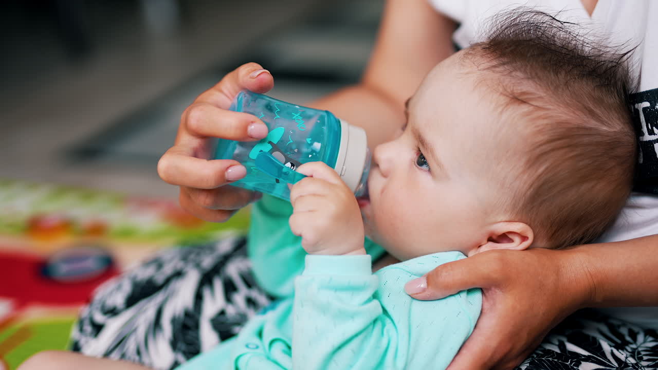 Cute boy half-sitting in mom's hands drinks water. Adorable baby with funny hair suckling bottle.
