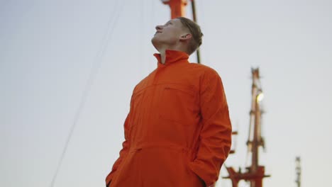 Apuesto-Joven-Trabajador-De-Almacén-De-Contenedores-Con-Uniforme-Naranja-Parado-Junto-Al-Barco-En-El-Puerto-Y-Mirando-Hacia-Arriba