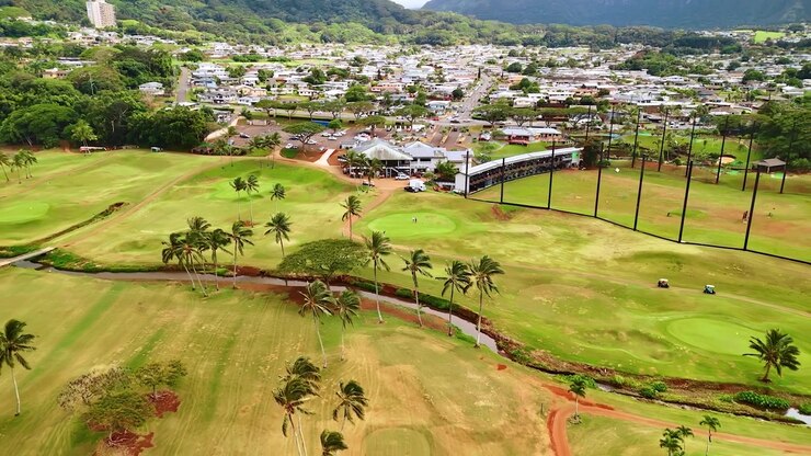 Palmen wachsen über dem Bach, der durch den grünen Golfplatz fließt. Einige Leute spielen Golf auf dem Platz. Stadtgebiet im Hintergrund. Hawaii, USA. Luftaufnahme