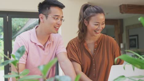 smiling asian couple sitting at table at home reviewing domestic finances using laptop