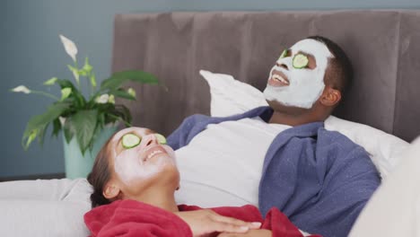 Video-De-Una-Feliz-Pareja-Diversa-Usando-Mascarillas-Limpiadoras-Y-Pepino-En-Los-Ojos-Acostados-En-La-Cama-Sonriendo