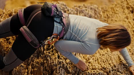 Vertical-video-of-a-blonde-girl-with-a-bob-hairstyle-in-a-gray-T-shirt-in-the-necessary-equipment-for-cleaving-and-using-a-safety-net,-climbs-up-a-yellow-rock