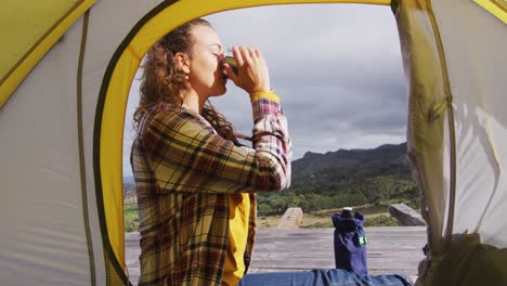 happy caucasian woman camping, sitting outside tent drinking coffee in rural mountain setting