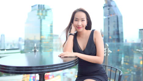 작은 뒷 드레스를 입은 젊은 여성이 옥상 바의 테이블에 앉아 있습니다.