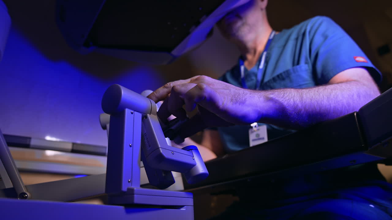 Doctor in blue uniform sits in front of operational system of Da Vinci equipment. Medic moves the handles and looks at special binoculars. Low angle view.