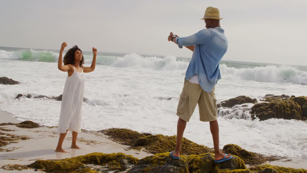 vista trasera de un hombre afroamericano haciendo clic en fotos de una mujer con teléfono móvil en la playa 4k