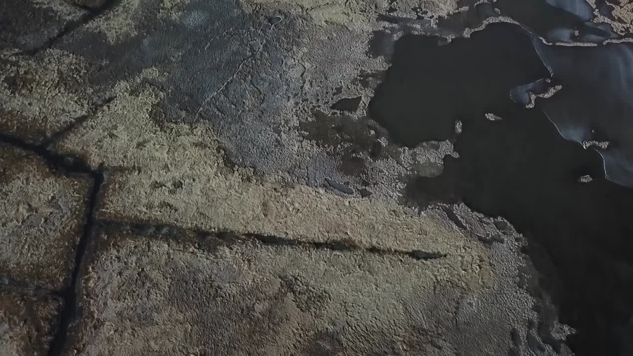 Top-down drone view of salt marsh pannes and tidal pools in the Great Marsh near Essex Bay, Massachusetts, dark water with thin ice edges and textured marsh grass, winter