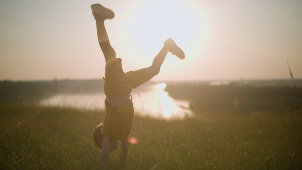 A young boy, dressed in yellow and wearing glasses, is caught mid-flip in a grassy field during a stunning sunset. The scene captures his playful energy, with a serene lake visible in the background