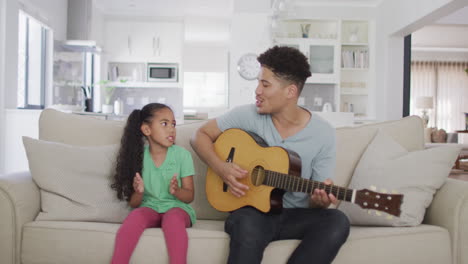 happy biracial father and daughter sitting on sofa playing guitar