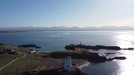 Vista-Aérea-Sobre-El-Idílico-Ynys-Llanddwyn-Con-La-Brumosa-Cordillera-De-Snowdonia-A-Través-Del-Brillante-Mar-Irlandés-Al-Amanecer.