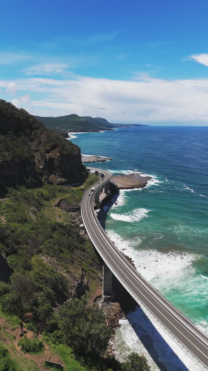 Driving cars on coastal road during sunny day with blue sky. Vertical drone wide shot. Waves of ocean. Traffic on Sea Cliff Bridge on Lawrence Hargrave to Wollongong,