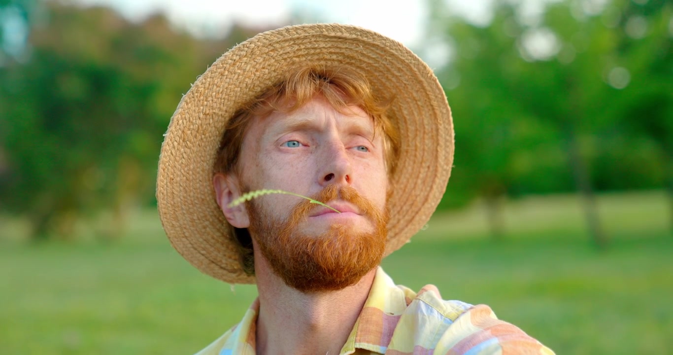 Relaxed Red-Bearded Man in Straw Hat Chewing Grass Outdoors