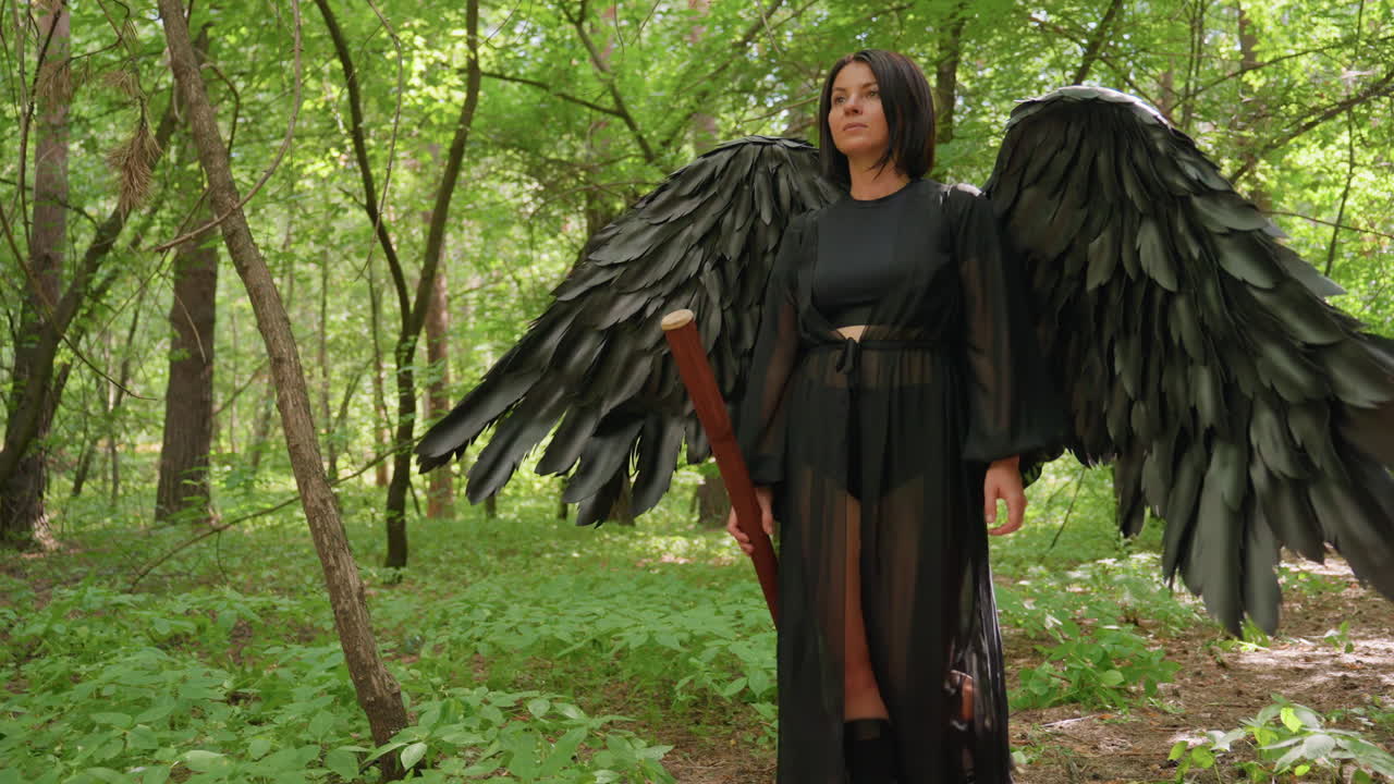 Goddess with large black wings walks gracefully through forbidden forest holding staff, sunlight filtering through leaves, blending elegance, power, and mystery in surreal atmosphere