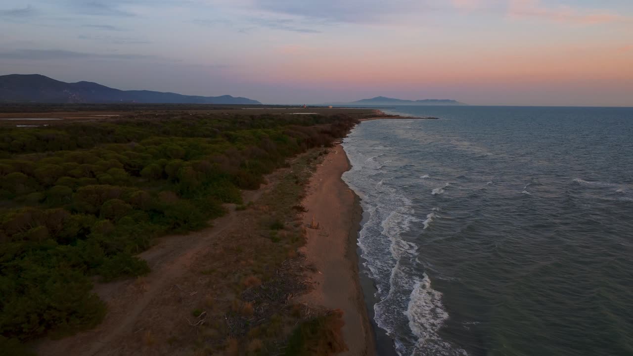 Riprese aeree cinematografiche ad alta quota con drone sopra una spiaggia sabbiosa al tramonto sul lungomare vicino a Castiglione, nell'iconico parco naturale della Maremma in Toscana, Italia, con onde, isole e un cielo rosso drammatico.