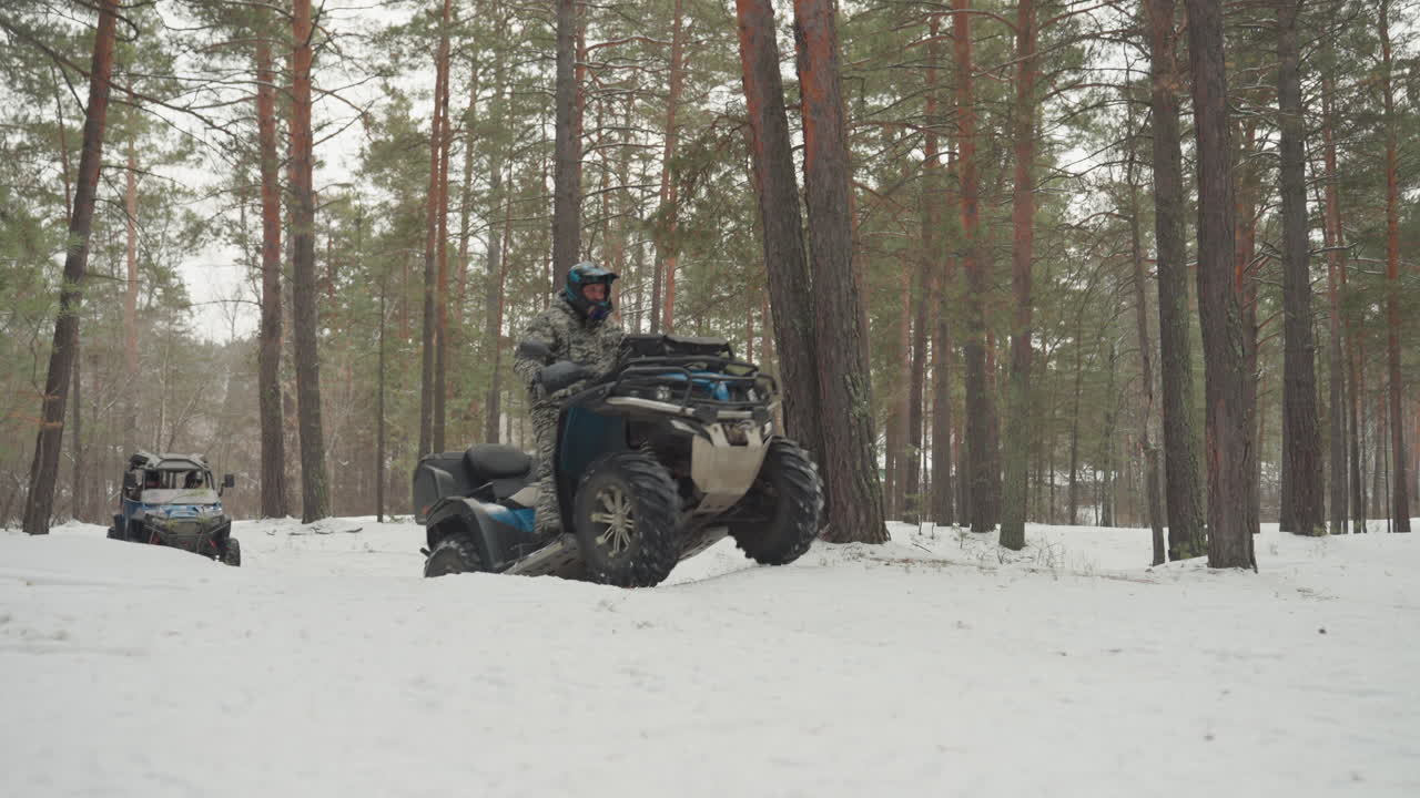Pilota di ATV in pineta innevata che naviga tra alberi alti, pilota camuffato su veicolo fuoristrada blu si muove lentamente sulla neve soffice, tracce di pneumatici e vapore di scarico visibili, luce nuvolosa, silenzio.