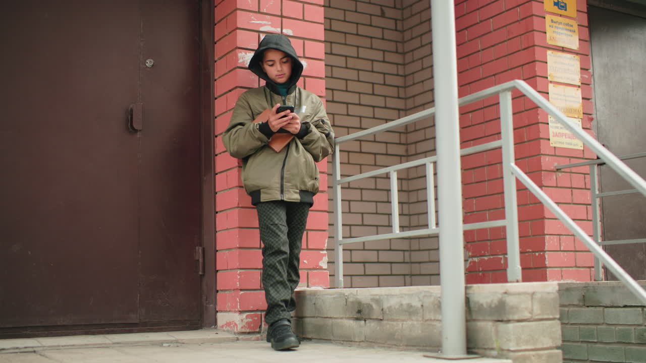 Teen in hooded jacket leans on building wall outdoors operating smartphone while holding brown book, focused on screen, standing near stair railing against red brick background