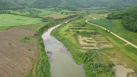 aerial footage tilting from a gorgeous landscape down to the canal and farmland, dirt road on the right, muak klek, saraburi, thailand
