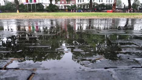 Detailed-shot-of-the-rain-falling-on-the-asphalt-while-people-walk-in-background